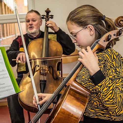 Jugendliche spielt konzentriert Cello vor einem Notenständer, im Hintergrund der Cello spielende Lehrer