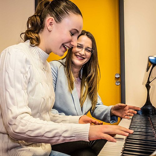 Junge Teenagerin spielt freudig Flügel, die Musiklehrerin sitzt daneben und gibt eine Anweisung