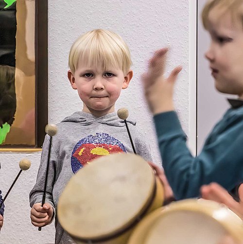 Zwei Vorschulkinder spielen auf Rahmentrommeln, ein weiteres schwingt im Hintergrund Schlegel für ein Xylophon