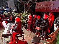 Geschwister spielen vierhändig am E-Piano, weitere Mitwirkende warten auf ihren Auftritt.