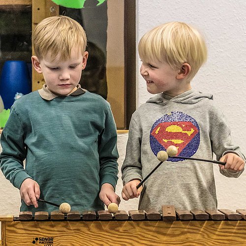 Zwei Vorschulkinder spielen auf einem Xylophon, eines ganz konzentriert, das andere lacht das erste ausgelassen an